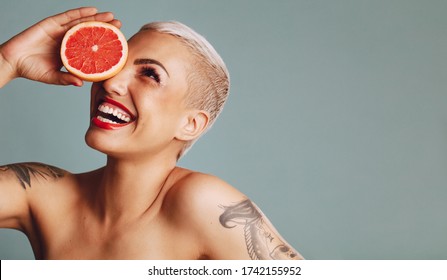 Close-up of a woman holding a grapefruit infront of her eye against grey background. Beautiful female model with short blond hair holding a grapefruit slice and smiling.