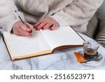 Close-up of a woman holding a pen, writing something in a notepad, working at a marble table and drinking coffee outside in a cafe during the cold season. A woman writes in her diary while working  