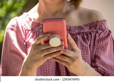 Close-up Of Woman Holding And Reading From A Cell Phone In Pink Case With Grip Handle On Back  -  Off Shoulder Blouse And Short Hair - Selective Focus And Unrecognizable