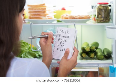 Close-up Of Woman Holding Spiral Book With Check List Against Open Refrigerator At Home