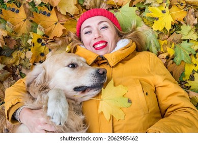 Close-up of a woman in a jacket and a retriever dog lie among the yellow autumn leaves in the park.