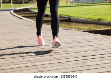 Closeup Of Woman Jogging In City Park. Lady Wearing Sports Pants, Sneakers And Training On Wooden Floor With Green Lawn In Background. Workout Concept.