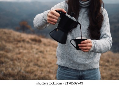 Close-up Of A Woman Pouring Out Coffee From A Moka Pot Into A Cup Outdoors In Autumn Time. Girl In A Cozy Casual Sweater In Front Of Mountains