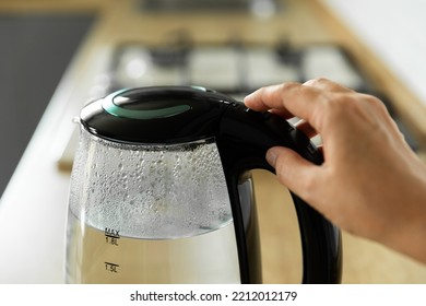 Close-up Of A Woman Pressing The Power Switch On An Electric Kettle.Save Energy At Home.A Modern Electric Transparent Kettle On A Wooden Table In The Kitchen. A Kettle For Boiling Water And Making Tea