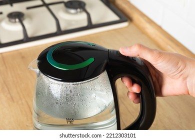 Close-up Of A Woman Pressing The Power Switch On An Electric Kettle.Save Energy At Home.A Modern Electric Transparent Kettle On A Wooden Table In The Kitchen. A Kettle For Boiling Water And Making Tea