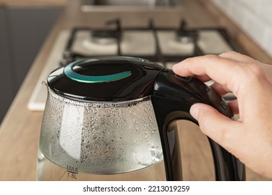 Close-up Of A Woman Pressing The Power Switch On An Electric Kettle.Save Energy At Home.A Modern Electric Transparent Kettle On A Wooden Table In The Kitchen. A Kettle For Boiling Water And Making Tea