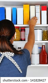 Closeup of a woman reaching into her pantry for a box of cereal. The well stocked cabinet is full of canned food, boxes, and bottles of typical grocery items. Items have blank labels.