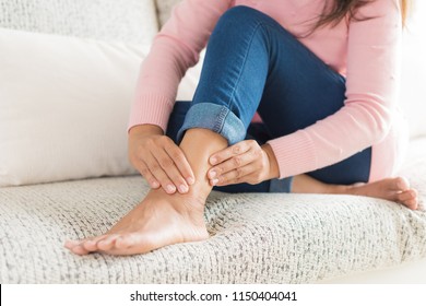 Closeup woman sitting on sofa holds her ankle injury, feeling pain. Health care and medical concept.