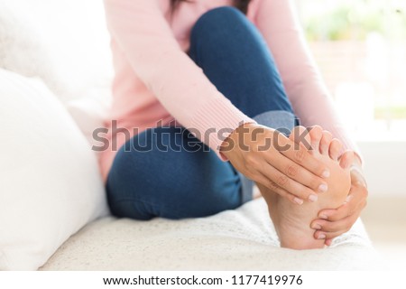 Closeup woman sitting on sofa holds her foot injury, feeling pain. Health care and medical concept.