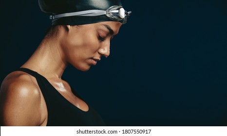 Close-up Of A Woman Swimmer Looking Tired After A Race. Female Wearing Black Swimsuit, Swimming Cap And Goggles On Dark Background.