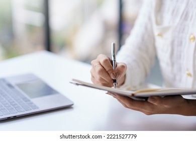 Close-up of a woman taking notes on notebook with laptop in office.