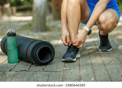 Close-up of a woman tying her shoes next to a yoga mat and water bottle, preparing for an outdoor workout session. - Powered by Shutterstock