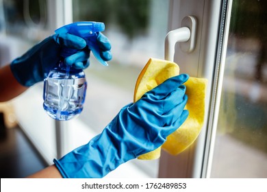Closeup of woman wearing blue rubber gloves cleaning the window handle with microfiber rag and sanitizing spray during coronavirus pandemic quarantine. Health care and adaptation concept.
