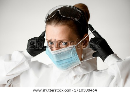 Close-up woman in white medical protective clothing and black latex gloves on her hands putting on mask on her face. White background.