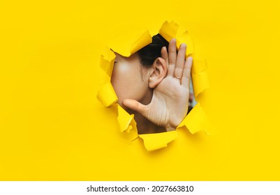Close-up of a woman's ear and hand through a torn hole in the paper. Yellow background, copy space. The concept of eavesdropping, espionage, gossip and tabloids.