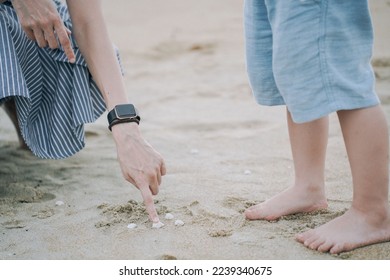 A closeup of a woman's finger touching tiny seashells on the sand next to a little boy's legs