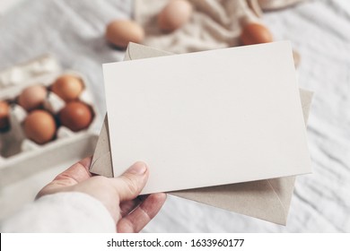 Closeup Of Woman's Hand Holding Blank Paper Card. Greeting Card Mock-up Scene. Spring, Easter Design. Feminine Styled Stock Photo. Blurred Background With Chicken Eggs And Line Tablecloth. Top View.