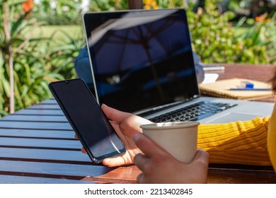 Close-up Of A Woman's Hand Holding A Mobile Phone On A Wooden Table Outside And A Blurred Laptop In The Background