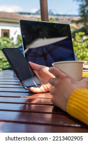 Close-up Of A Woman's Hand Holding A Mobile Phone On A Wooden Table Outside And A Blurred Laptop In The Background