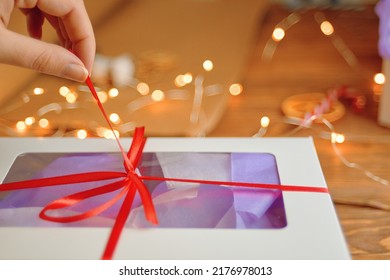 Close-up Of Woman's Hand Pulling Ribbon From Surprise Box. Unpacking Gift On Wooden Table. Bokeh Lights In Background. Festive Concept Of Christmas Or Birthday.