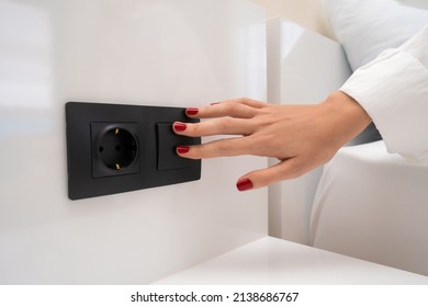 Close-up of a woman's hand with red nail polish turns on the light near the bed with a black switch on a white wall, next to the outlet - Powered by Shutterstock