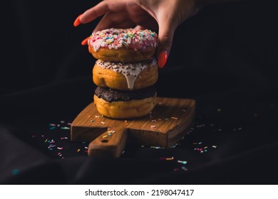 Close-up of a woman's hand taking a delicious donut from a wooden board. Delicious donuts laid out on a wooden cutting board on a black background.