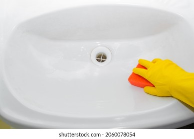 Close-up of woman's hand in yellow rubber glove with red sponge washes and polishes washbasin in bathroom indoors, top view. Regular general cleaning and bath hygiene.