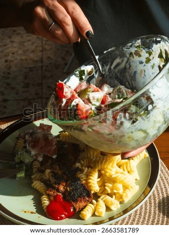 Close-up of a woman's hands adding a portion of fresh cucumber and tomato salad with yogurt dressing to a plate already containing fusilli pasta and grilled meat with ketchup
