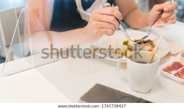 Closeup of woman\'s hands eating alone with medical face\
mask hang on her neck sit on other side of acrylic divider /\
barrier on table. New normal & Social distancing during\
Covid-19 pandemic 