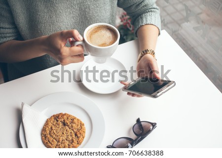 Close-up of woman's hands holding cell phone while drinking coffee and eating oat cookie in modern cafe. Reading news, social network, surfing the web / internet in smartphone. Coffee break business.