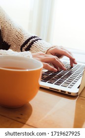 closeup of woman's hands typing on a laptop keyboard with orange mug containing tea on the wood rustic background desk. concept of home office and distance working during coronavirus lockdown at home
