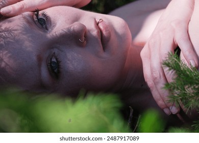 close-up of a woman's serene face lying among green foliage, with delicate shadows cast by the leaves. Her expression is calm and introspective, capturing a moment of peaceful connection with nature. - Powered by Shutterstock