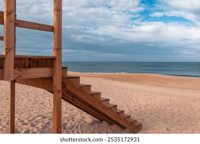 Close-up of wooden lifeguard tower stairs on a serene sandy beach in Portugal, with Atlantic Ocean in the background under a partly cloudy blue sky, Tranquil coastal landscape - Powered by Shutterstock