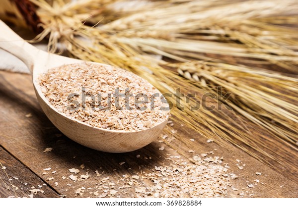 Close-up of wooden spoon filled with wheat bran on\
the background of wheat ears. Dietary supplement to improve\
digestion. Source of dietary fiber. Wooden planks background.\
Shallow depth of field