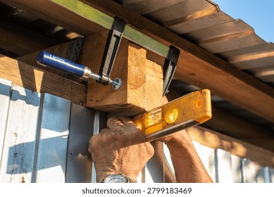 Close-up of worker hands using a yellow bubble spirit level and an F-clamp tool to align a wooden block during roof construction