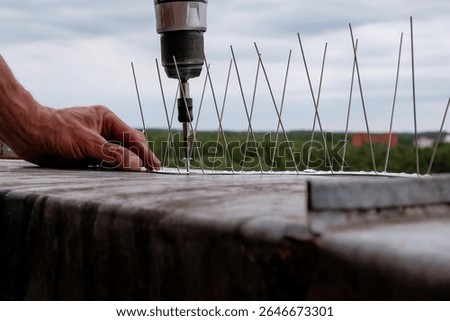 Close-up of a worker using a cordless power drill to install stainless steel bird deterrent spikes on a rooftop edge, with protective anti-pigeon barrier system in focus
