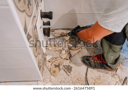 A close-up of a worker using a power tool to break bathroom floor tiles during renovation, with debris and broken ceramic pieces scattered.