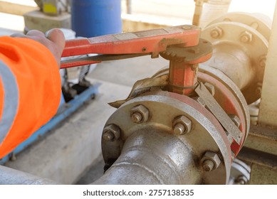 Close-up of a worker's gloved hand using a wrench to operate a red valve on a large industrial pipeline in a factory setting.