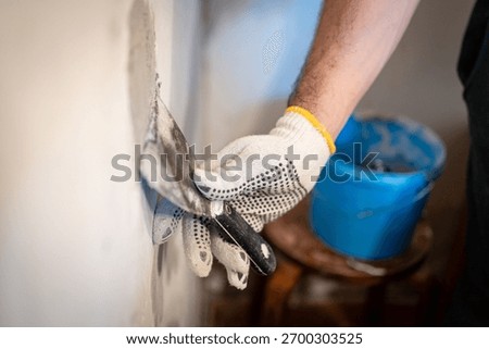 Close-up of a worker's gloved hand using a small putty knife to press thick patching compound firmly into a hole in the interior wall.