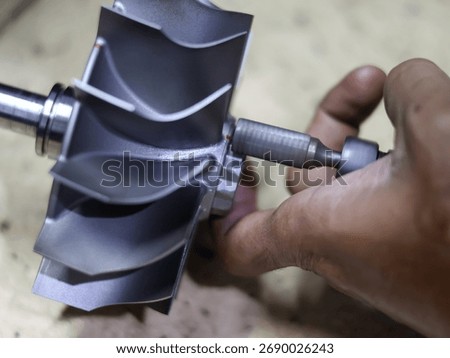 Close-up of a worker's hand using a bor tuner or borcun to perform a correction or balance adjustment on the metal blades of a turbocharger compressor wheel.