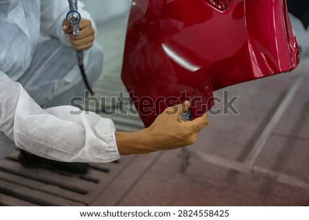 Close-up of a worker's hands removing protective film from a glossy red painted car bumper in an auto body shop.