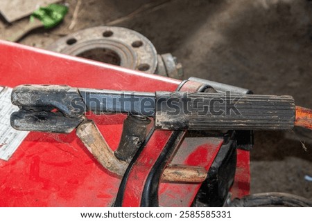 Close-up of a worn welding electrode holder (stang las) used in industrial metal fabrication. The handle and clamp show signs of heavy use, highlighting essential tools for welding, repairs