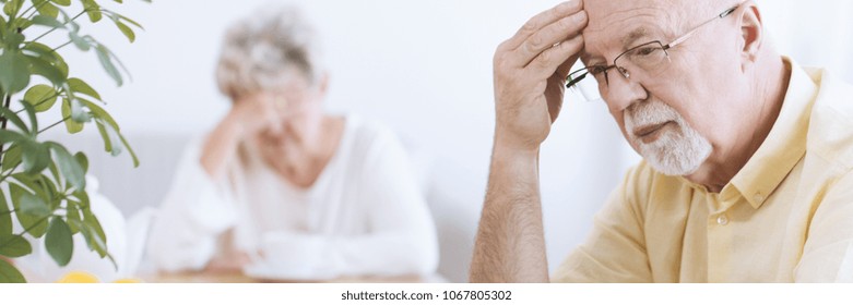 Close-up of a worried, elderly man deep in thoughts, with his hand on his forehead and a sad senior woman blurry in the background