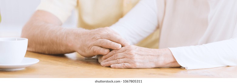 A close-up of wrinkled arms on a table, an older man holding his hand on an elderly woman's hands showing affection and supporting her - Powered by Shutterstock