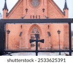 A close-up of a wrought iron fence with a cross design in front of a brick church building. The church has a visible date of 1863 and features gothic architectural elements.