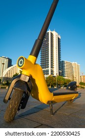 Close-up Of A Yellow Electric Scooter Stands On A Tile In A City Park Against The Background Of Modern Buildings At Sunset. Eco-friendly Mode Of Transport For The City. Vertical Photo