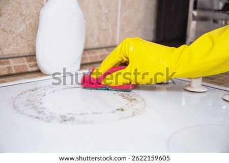 Close-up of yellow gloved hand using red sponge to clean dirty white stove ring in tiled kitchen. Cleaning, household chores