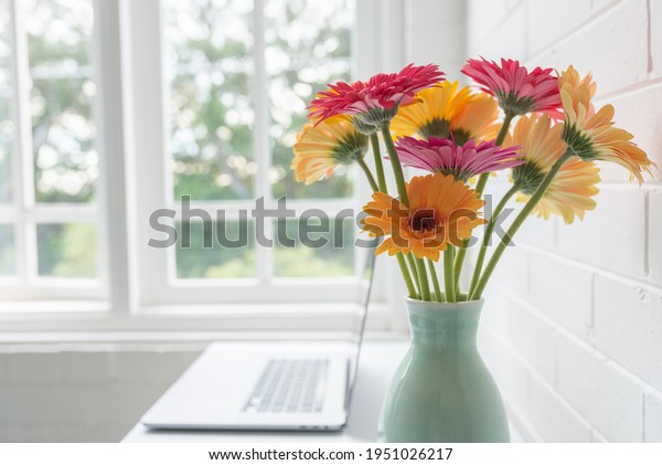 Closeup of yellow\
and pink gerbera daisies in green vase on desk with laptop next to\
window (selective\
focus)