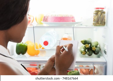 Close-up Of Young African Woman Writing On Spiral Book Near Open Refrigerator