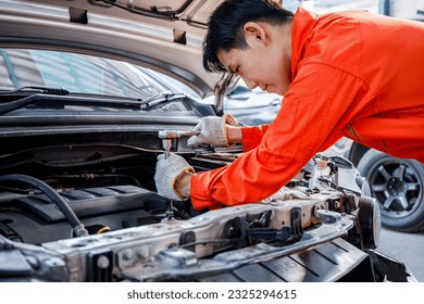 Close-up of a young Asian technician in an orange uniform. Car repairing wrench on car engine in the garage. Repair service. Skilled mechanic working behind the scenes with cars and car parts. - Shutterstock ID 2325294615
